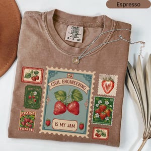 May include: Espresso-coloured t-shirt with a strawberry design. The shirt displays postage stamp-style graphics with strawberries and the text "CIVIL ENGINEERING IS MY JAM". A silver necklace with a pendant rests on the shirt.
