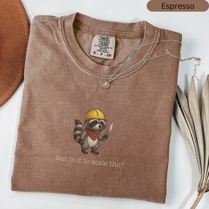 May include: Espresso-coloured t-shirt featuring a raccoon graphic wearing a yellow hard hat and holding a knife. The text "but is it to scale tho?" is below the graphic. A silver necklace with a pendant is draped across the shirt.