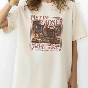 Op de afbeelding: Ivoor kleurige oversized t-shirt met de tekst "GET IN LOSER" boven een illustratie van een persoon die in bed leest. De tekst "WE'VE GOT ONE MORE CHAPTER TO READ" staat eronder. De woorden "midnight readers club" staan onderaan.