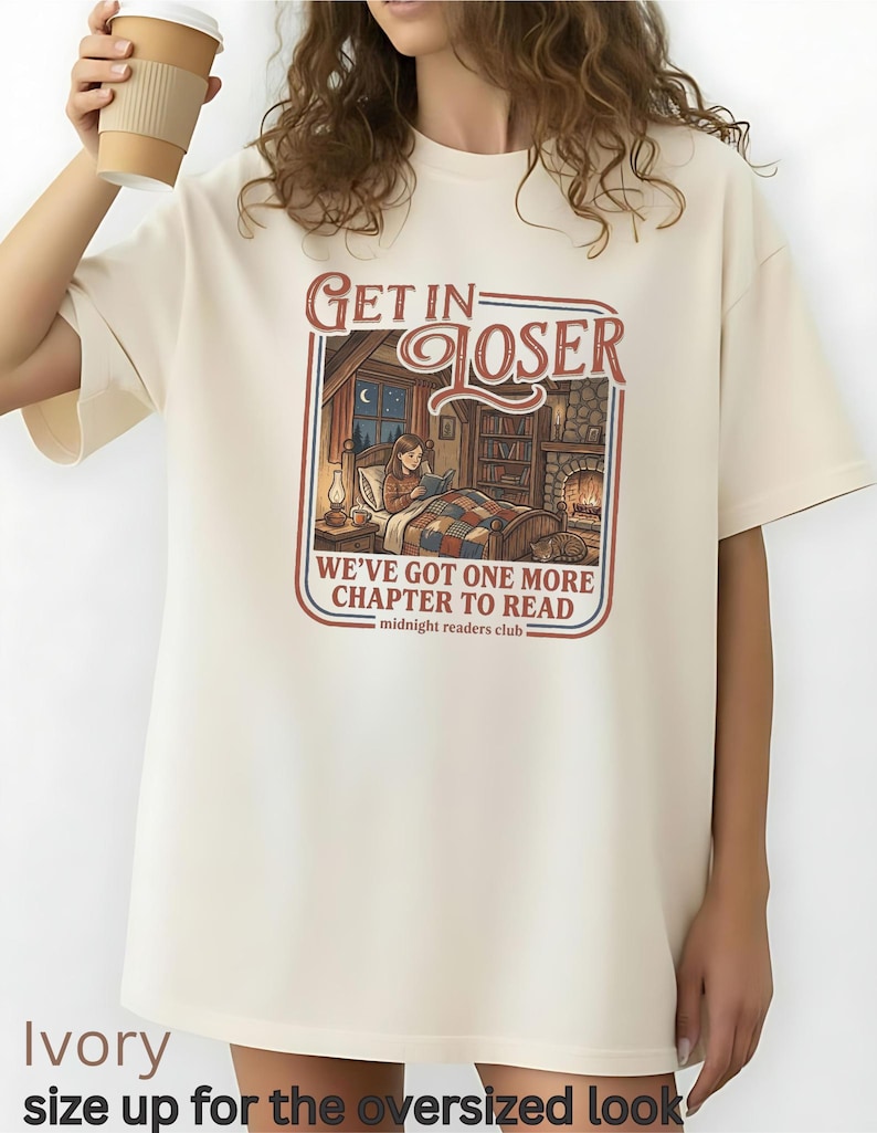 Op de afbeelding: Ivoor kleurige oversized t-shirt met de tekst "GET IN LOSER" boven een illustratie van een persoon die in bed leest. De tekst "WE'VE GOT ONE MORE CHAPTER TO READ" staat eronder. De woorden "midnight readers club" staan onderaan.