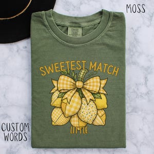 May include: A moss green t-shirt featuring a lemon and bow graphic. The text "Sweetest Match Little" is printed in yellow. The lemons have various yellow patterns. The shirt is made of a soft material.