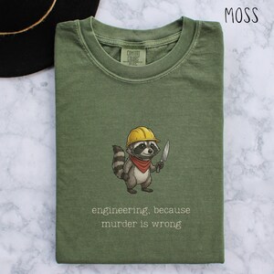 May include: Moss green t-shirt featuring a cartoon raccoon in a yellow hard hat and red bandana, holding a knife. The text reads "engineering, because murder is wrong". The shirt is made of Comfort Colors material.