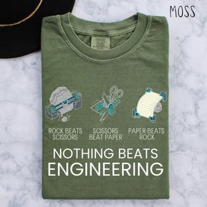 May include: Moss green t-shirt with the text "NOTHING BEATS ENGINEERING". The shirt features a graphic of rock, scissors, and paper with the phrases "ROCK BEATS SCISSORS", "SCISSORS BEAT PAPER", and "PAPER BEATS ROCK".