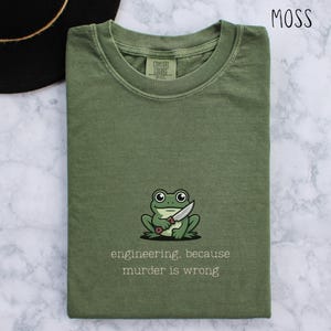 May include: Moss green t-shirt featuring a frog graphic with a knife. The text below the frog reads "engineering, because murder is wrong." The shirt is made of a soft material and has a crew neck.