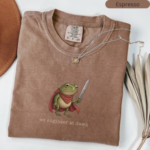 May include: Espresso-coloured t-shirt featuring a cartoon frog in a red cape, holding a sword, with the text "we engineer at dawn". A silver necklace with a pendant is draped across the shirt. The shirt is a Comfort Colors brand.