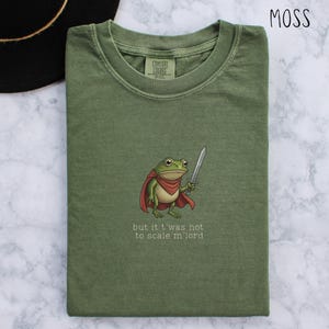 May include: Moss green t-shirt featuring a cartoon frog in a red cape, holding a sword. The text "but it t'was not to scale m'lord" is printed below the frog. The shirt is folded and has a Comfort Colors label.