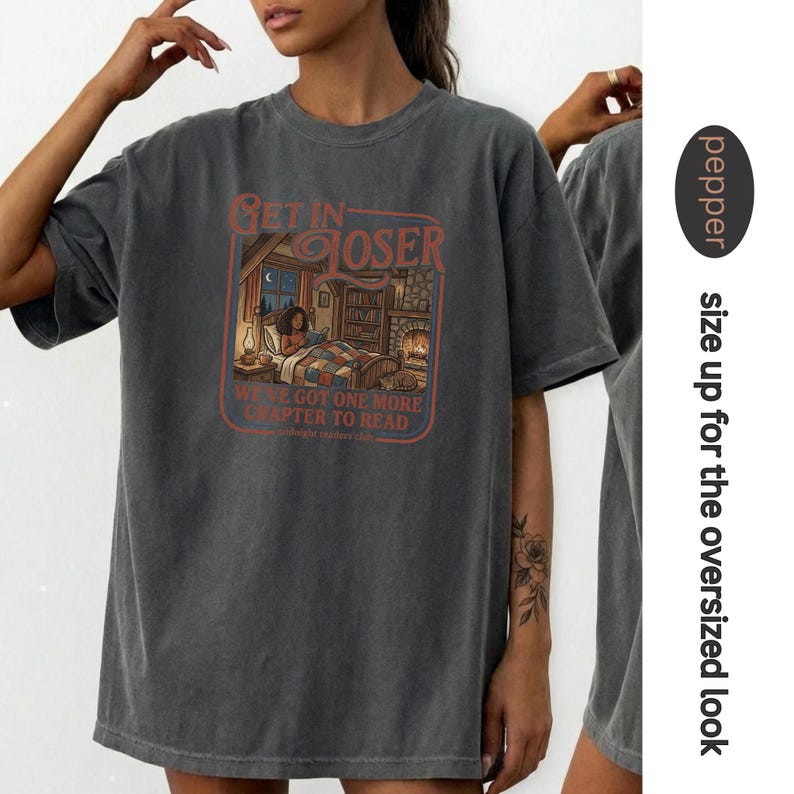 Op de afbeelding: Donkergrijs oversized T-shirt met de tekst "GET IN CLOSER" en een illustratie van een persoon die in bed leest. De tekst "WE'VE GOT ONE MORE CHAPTER TO READ" is ook inbegrepen. Het shirt is gestyled voor een oversized look.