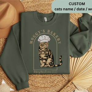 May include: Green sweatshirt with a cat wearing a chef's hat and the text "Moggy's Bakery Baking Biscuits since 2023".