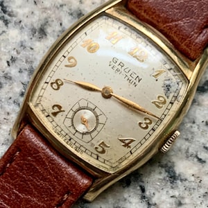 Op de afbeelding: Vintage Gruen Veri-Thin polshorloge met een goudkleurige rechthoekige kast en een crèmekleurige wijzerplaat. Het horloge heeft gouden cijfers en wijzers, een kleine secondenwijzer en een bruine leren band. Het horloge is gemarkeerd met "SWITZERLAND".