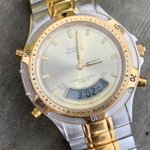May include: A two-tone gold and silver Armitron Pro-Air Sport watch with a digital display showing the time 3:02:02. The watch has a round face with a white background and black markings. The watch is water resistant to 100 meters.