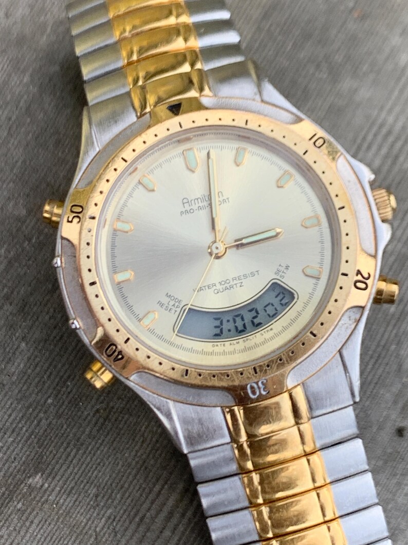 May include: A two-tone gold and silver Armitron Pro-Air Sport watch with a digital display showing the time 3:02:02. The watch has a round face with a white background and black markings. The watch is water resistant to 100 meters.