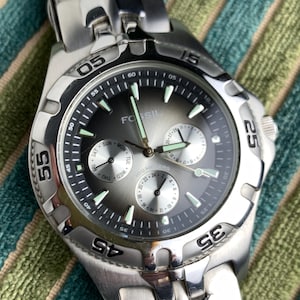 Peut inclure: Une montre Fossil en acier inoxydable avec un cadran gris et plusieurs sous-cadrans. La montre comporte des aiguilles et des marqueurs lumineux, ainsi qu'un affichage de la date. La lunette comporte des chiffres aux repères 5, 15, 25, 35, 45 et 55 minutes.