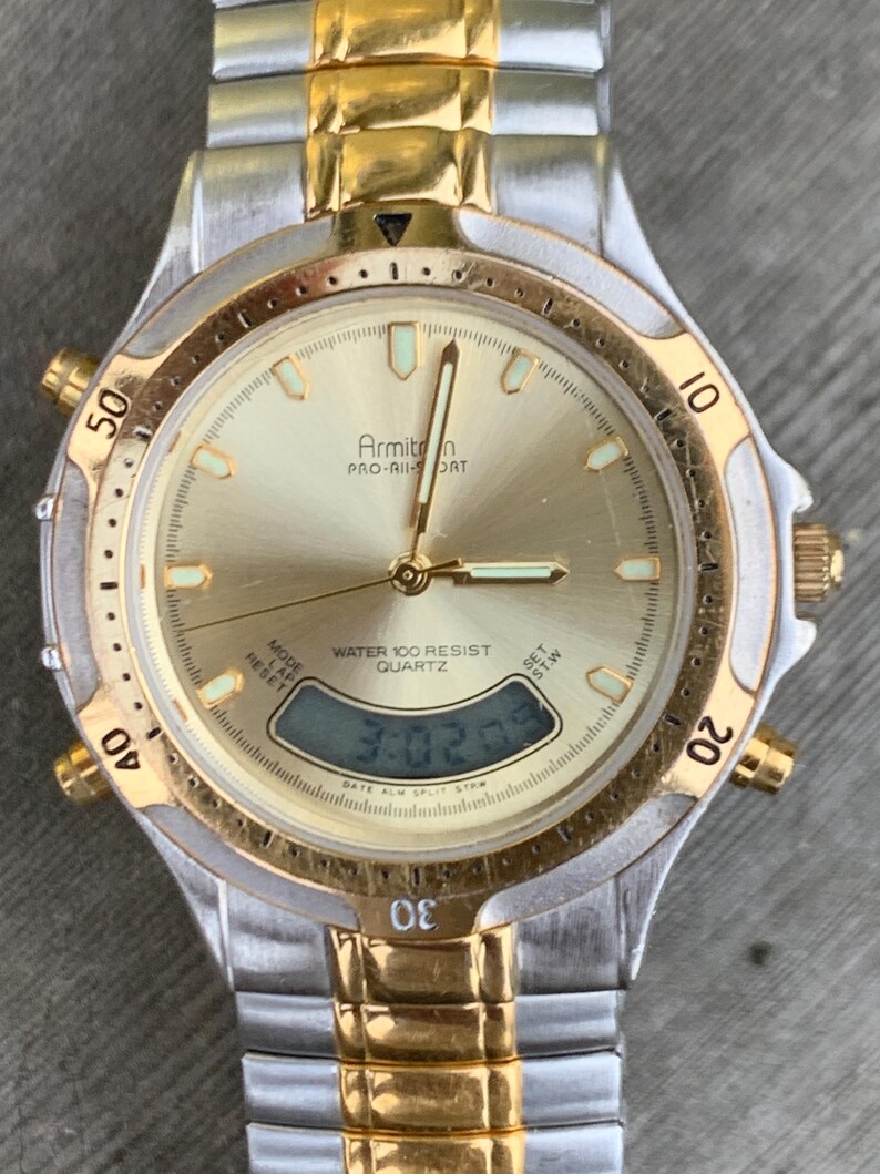 May include: A gold and silver two-tone Armitron Pro-Air Sport watch with a digital display and a gold-tone bezel. The watch face is gold with black markings and the text "Armitron Pro-Air Sport" and "Water 100 Resist Quartz".