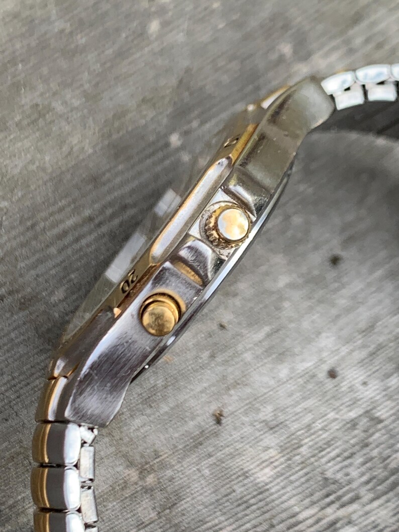 May include: A silver and gold wristwatch with a metal band. The watch has a round face and three gold buttons on the side.