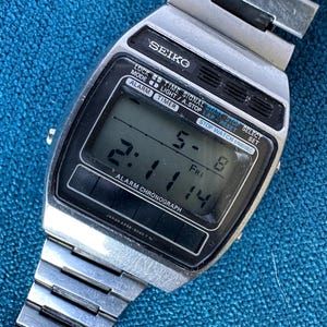 May include: A vintage Seiko digital watch with a silver metal strap and a black rectangular face. The watch displays the time, date, and various functions. The watch face has the text "SEIKO", "ALARM CHRONOGRAPH", and other function labels.