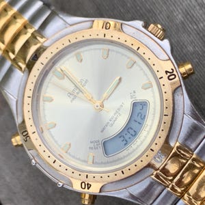 May include: A two-tone gold and silver analog and digital wristwatch with a white face. The watch has a date display, a chronograph, and a water resistance of 100 meters. The watch face has the text "Aim bn PAO A FORT QUARTZ WATER 100 RESIST" printed on it.