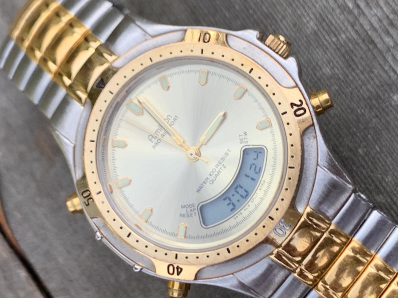 May include: A two-tone gold and silver analog and digital wristwatch with a white face. The watch has a date display, a chronograph, and a water resistance of 100 meters. The watch face has the text "Aim bn PAO A FORT QUARTZ WATER 100 RESIST" printed on it.