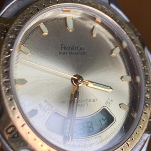 May include: A gold and silver Armitron Pro-All-Sport watch with a digital display and a gold-tone bezel. The watch face is white with black markings and the text "WATER RESIST QUARTZ" is printed on the face.