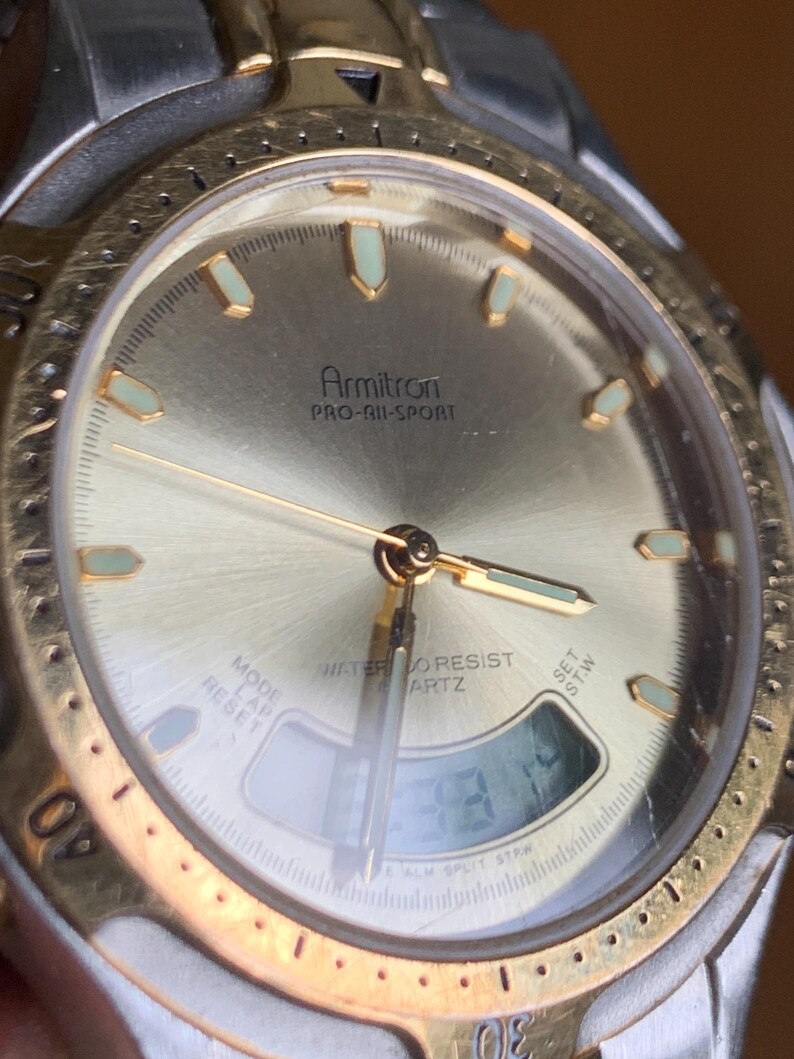 May include: A gold and silver Armitron Pro-All-Sport watch with a digital display and a gold-tone bezel. The watch face is white with black markings and the text "WATER RESIST QUARTZ" is printed on the face.