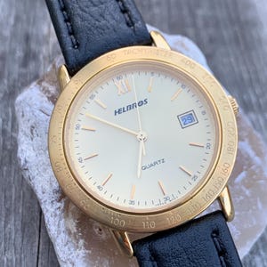 Könnte beinhalten: Eine goldfarbene Helbros-Quarzuhr mit cremefarbenem Zifferblatt, schwarzem Lederarmband und Datumsanzeige. Die Uhr hat römische Ziffern und eine Tachymeterskala. Die Uhr liegt auf einem hellen Stein.