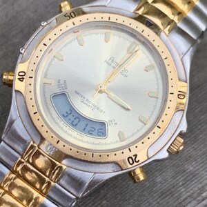May include: A silver and gold two-tone wristwatch with a white face and black digital display. The watch face has the words "Armon Pro-Fort" and "Water Resist Quartz".