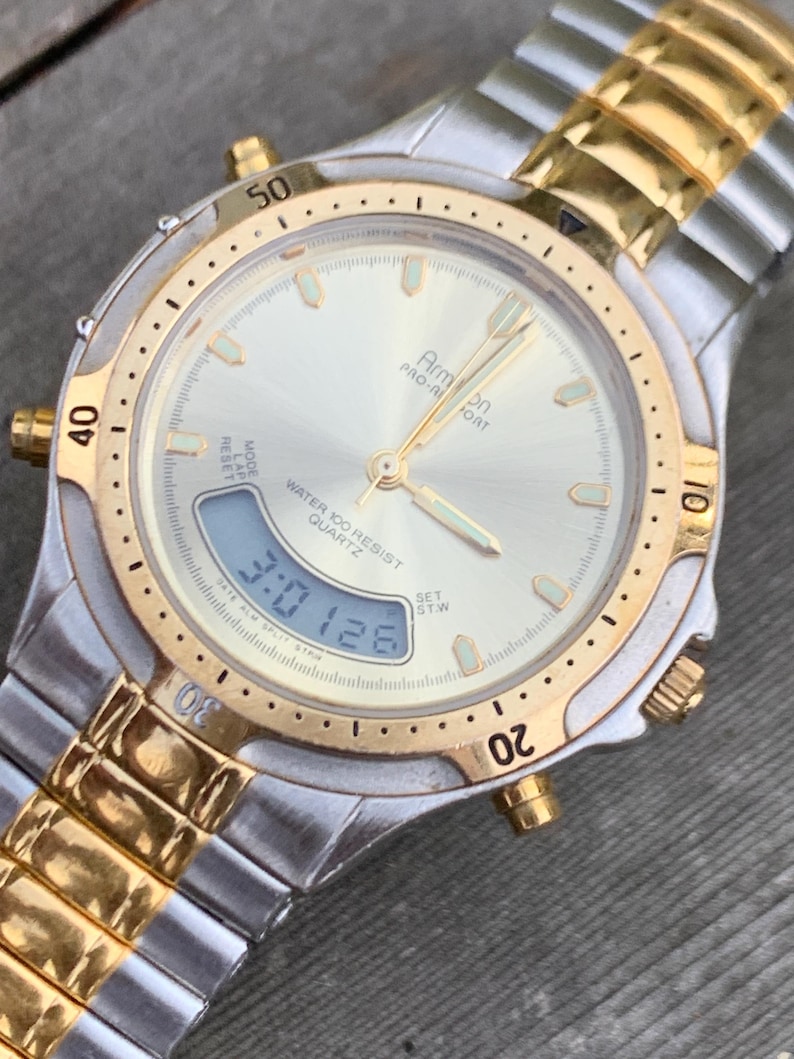 May include: A silver and gold two-tone wristwatch with a white face and black digital display. The watch face has the words "Armon Pro-Fort" and "Water Resist Quartz".