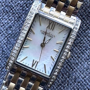 Op de afbeelding: Een rechthoekig Caravelle New York polshorloge met een parelmoeren wijzerplaat, gouden wijzers en Romeinse cijfers. Het horloge heeft een zilver-gouden band en is omlijst met een rij kleine, heldere kristallen.