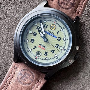 Könnte beinhalten: Eine schwarze Timex Expedition-Uhr mit hellem Zifferblatt und braunem Lederarmband. Das Zifferblatt zeigt die Uhrzeit, mit der Indiglo-Funktion. Das Armband hat ein gestempeltes Logo in der Nähe des Uhrengehäuses.