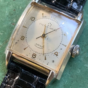May include: A silver-toned wristwatch with a white face and black leather band. The watch face has black Roman numeral hour markers and the text "ETERNA-MATIC" and "HIRKS CHALLENGER" printed on it.