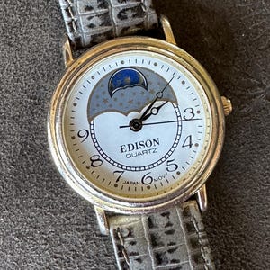 Peut inclure: Une montre Edison Quartz dorée avec un cadran blanc orné d'un motif de phases lunaires. La montre a des aiguilles noires et un bracelet en cuir gris avec un motif reptile. Le cadran affiche les chiffres 3, 4, 5, 6, 7 et 8.