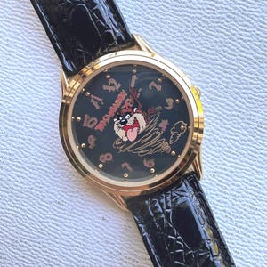 Könnte beinhalten: Goldfarbene Armbanduhr mit schwarzem Zifferblatt mit der Cartoonfigur Tasmanian Devil. Die Uhr hat ein schwarzes Lederarmband und roségoldfarbene Ziffern. Auf dem Zifferblatt steht auch das Wort "Tasmanian".