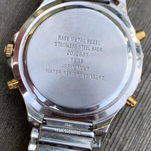 May include: The back of a silver and gold watch with the text "BASE METAL BEZEL STAINLESS STEEL BACK 20/2887 T205 JAPAN MOVT WATER RESISTANT 100FT."