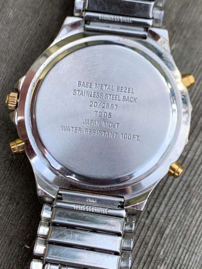 May include: The back of a silver and gold watch with the text "BASE METAL BEZEL STAINLESS STEEL BACK 20/2887 T205 JAPAN MOVT WATER RESISTANT 100FT."