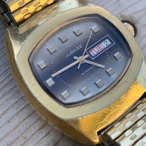 May include: A gold-toned wristwatch with a square face and a gold metal band. The watch face has a silver background with black hands and black Roman numeral hour markers. The watch face also has a small window that displays the day of the week, which is "TUE 2". The watch face also has the word "JUBILEE" and "AUTOMATIC" printed on it.