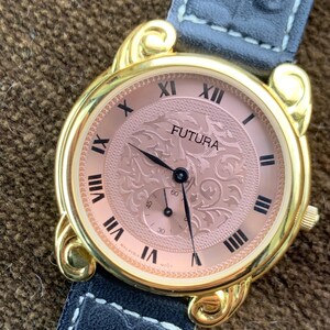 May include: A gold-toned Futura watch with a rose-colored face and black Roman numerals. The watch features a decorative engraved design on the face and a small subdial. It has a gray leather band.