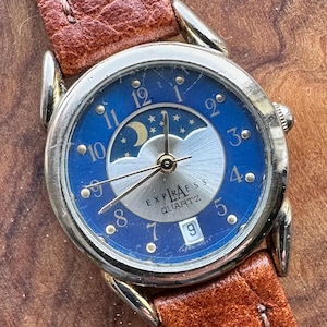 Peut inclure: Une montre-bracelet Express Quartz vintage avec un cadran bleu orné d'un motif de lune et d'étoiles. La montre a des accents dorés, un guichet de date et un bracelet en cuir marron.