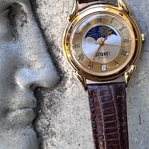 Puede incluir: Un reloj de pulsera dorado con una correa de cuero marrón texturizada. La esfera redonda presenta un fondo dorado con números romanos negros, un indicador de fase lunar y solar en la parte superior y una ventana de fecha con Gitano Quartz.