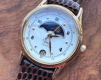 Könnte beinhalten: Eine goldfarbene Armbanduhr mit weißem Zifferblatt, Mondphasenanzeige, Datumsfenster mit der Zahl 24 und schwarzen arabischen Ziffern. Die Uhr hat ein braunes, strukturiertes Lederarmband und den Aufdruck Time Quartz auf dem Zifferblatt.
