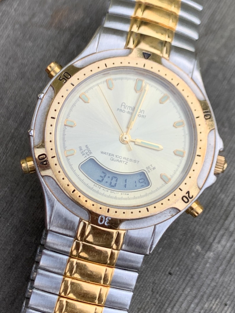 May include: A gold and silver two-tone Armilon Pro-Air Sport watch with a white face and a digital display. The watch is water resistant to 100 meters.