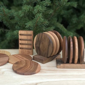 May include: Wooden coaster sets in various shades of brown. The image shows several round coasters, some stacked in holders, and a rectangular holder. The coasters have visible wood grain patterns, suggesting a natural finish. The background is a blurred green.