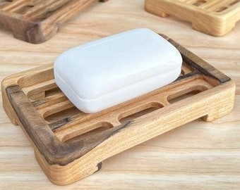 May include: Three wooden soap dishes, two are empty and one has a white bar of soap on top. The dishes are made of light and dark wood and have a slatted design.