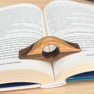 May include: A wooden book holder with a dark brown stain and a natural wood finish. The holder is shaped like a bridge and has a hole in the center for the book page. The holder is resting on a book with the text "PER PALACE by Miranda Cowley Heller" visible.