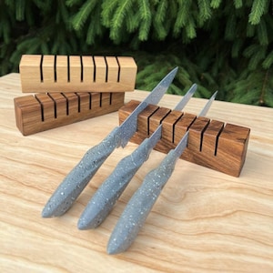 Puede incluir: Un bloque de cuchillos de madera con tres cuchillos grises moteados insertados. El bloque está hecho de madera oscura y tiene ocho ranuras para cuchillos. El bloque está sobre una superficie de madera clara.