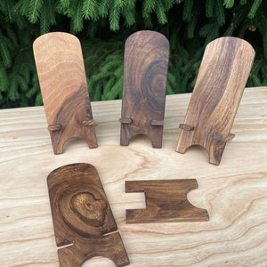 Puede incluir: Cinco soportes de teléfono de madera en varios tonos de marrón. Los soportes están hechos de madera maciza y tienen un diseño simple y minimalista.