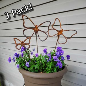 May include: Three rusty metal butterfly garden stakes are displayed in a brown flower pot filled with purple pansies. The text "3 Pack!" is in the upper left corner. The butterflies have openwork wings.