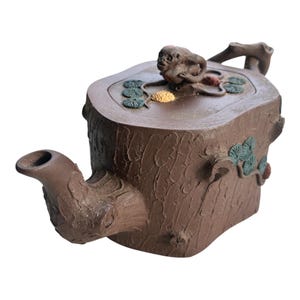Puede incluir: Una tetera de arcilla marrón con forma de tocón de árbol y superficie de corteza texturizada. La tapa presenta una figura de mono esculpida, pequeños grupos de agujas de pino verdes y un detalle amarillo. Un pico curvo sobresale.