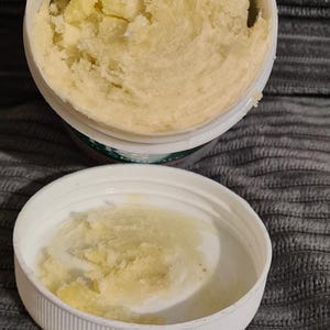 May include: Open white jar filled with a light yellow, creamy substance. The lid is next to the jar, also containing some of the substance. The jar is on a gray, textured surface.