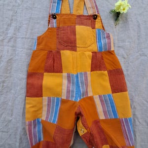 May include: A patchwork baby romper in shades of orange, yellow, and blue. The garment features a bib front with a pocket, adjustable straps, and snap closures at the legs. The fabric is a mix of solid and striped patterns. A small bouquet of yellow flowers rests nearby.