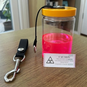 Puede incluir: Un frasco de vidrio transparente con tapa amarilla y asa de correa negra. El frasco contiene un líquido rosa brillante. Una etiqueta de advertencia en la parte delantera del frasco dice: "¡ATENCIÓN! -CARGADO NEGATIVAMENTE- PSYCHOMAGNOTHERIC SLIME 'Slime de humor' ¡Responde a la emoción humana!"