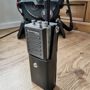 Puede incluir: Una funda de radio de cuero negro con una radio Motorola MT 500 plateada en su interior. La radio tiene una antena negra y tres botones plateados.